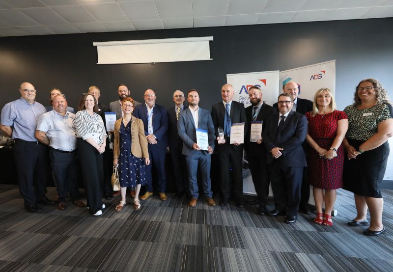 Group of people in business attire holding awards and certificates at an ADS and Supply Chain Solutions event with branded banners in the background.