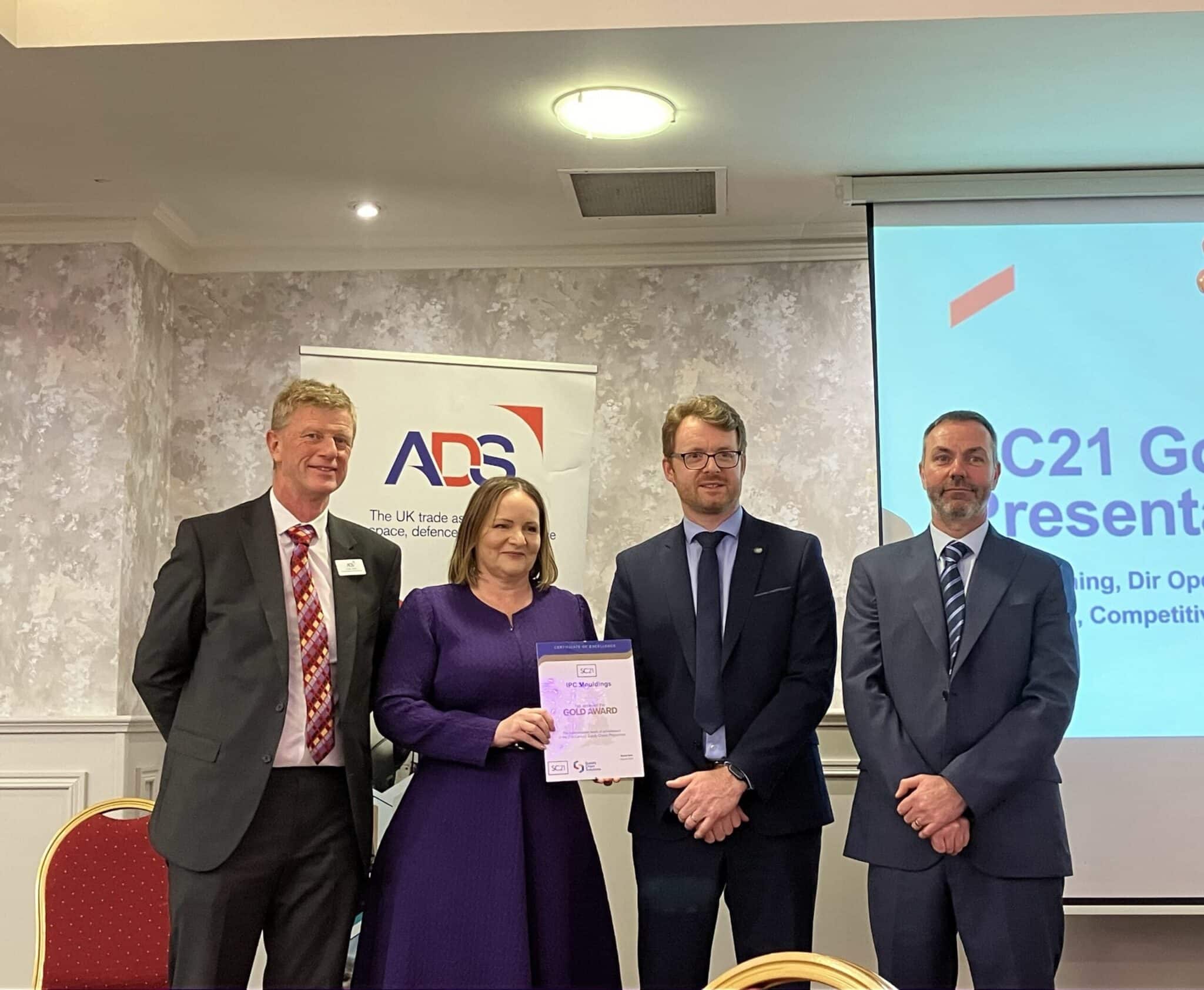 Four people at an ADS event stand together as a woman holds the SC21 Gold Award certificate for IPC Mouldings, with a presentation screen and banner in the background.