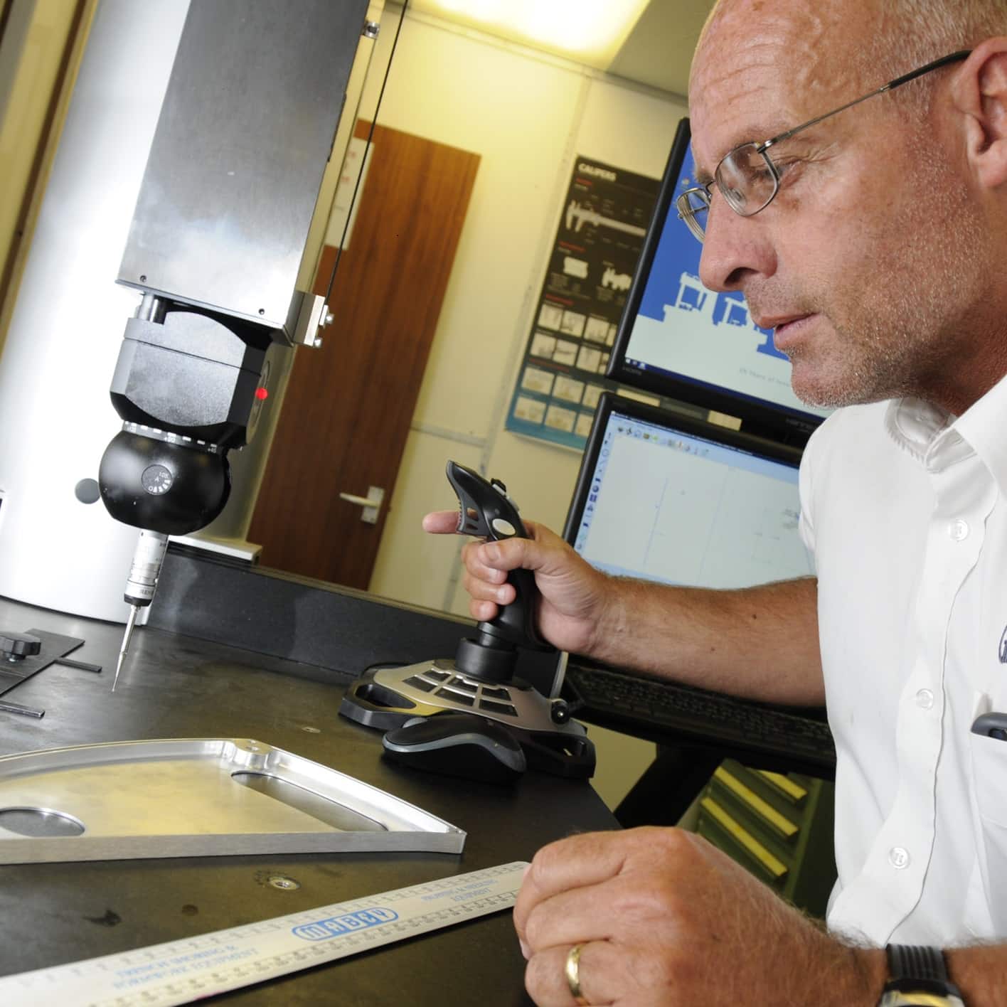 Engineer using a joystick to operate a coordinate measuring machine (CMM) for precise inspection of a machined metal component in a quality control lab.