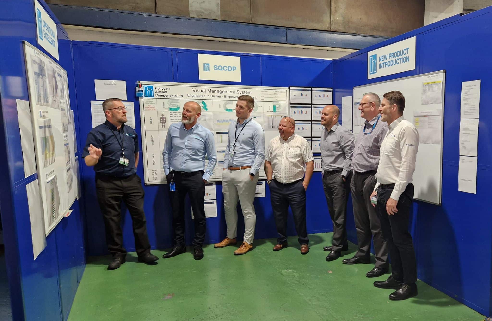 A team of seven men at Hollygate Aircraft Components Ltd stand in front of a Visual Management System display, engaging in a team briefing in a blue-walled workspace.