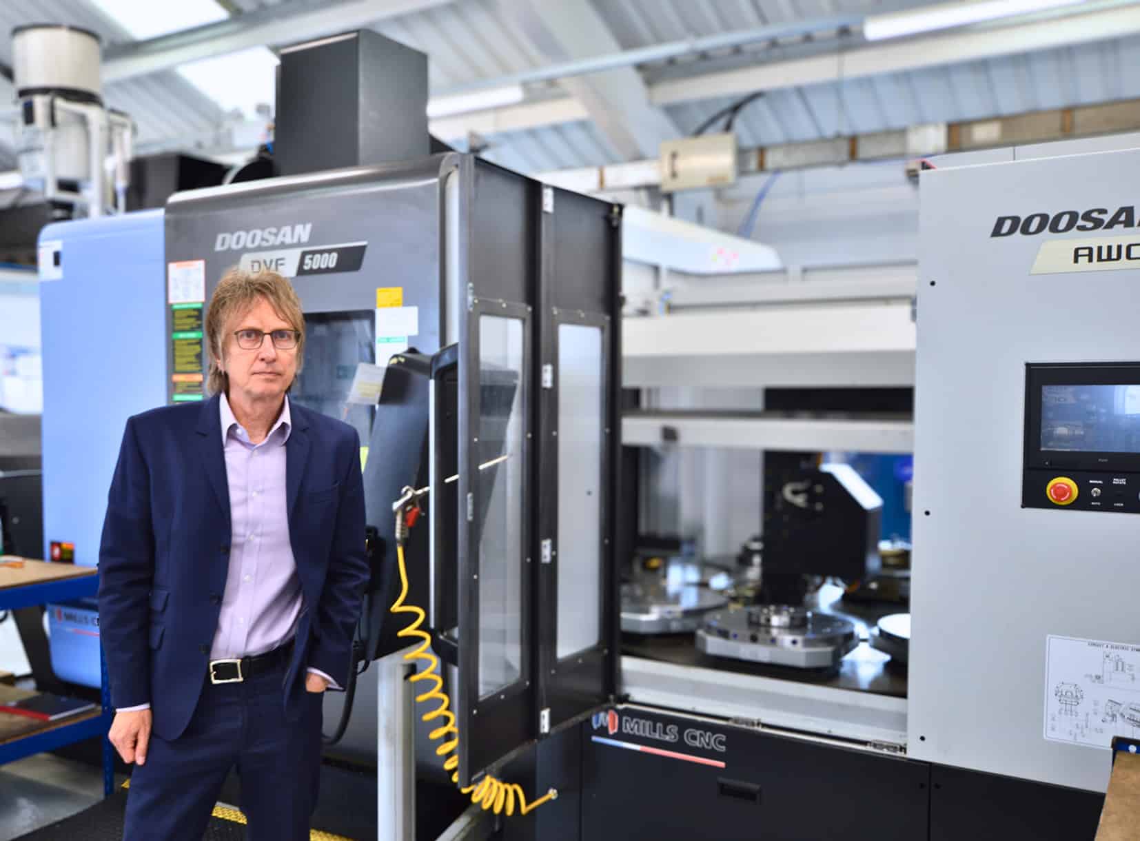 Man in a suit standing beside a Doosan DVF 5000 CNC machine inside a high-tech manufacturing facility, showcasing advanced automation at Mills CNC.
