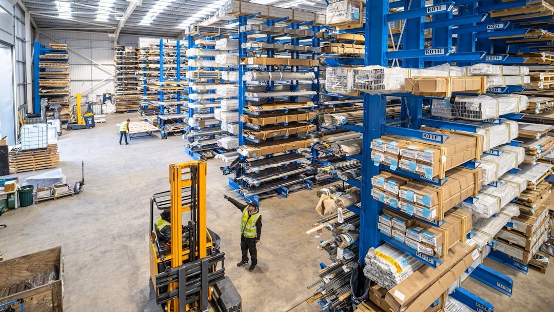 Interior of a large warehouse storing aluminium profiles on blue cantilever racking, with workers operating a forklift and handling stock.