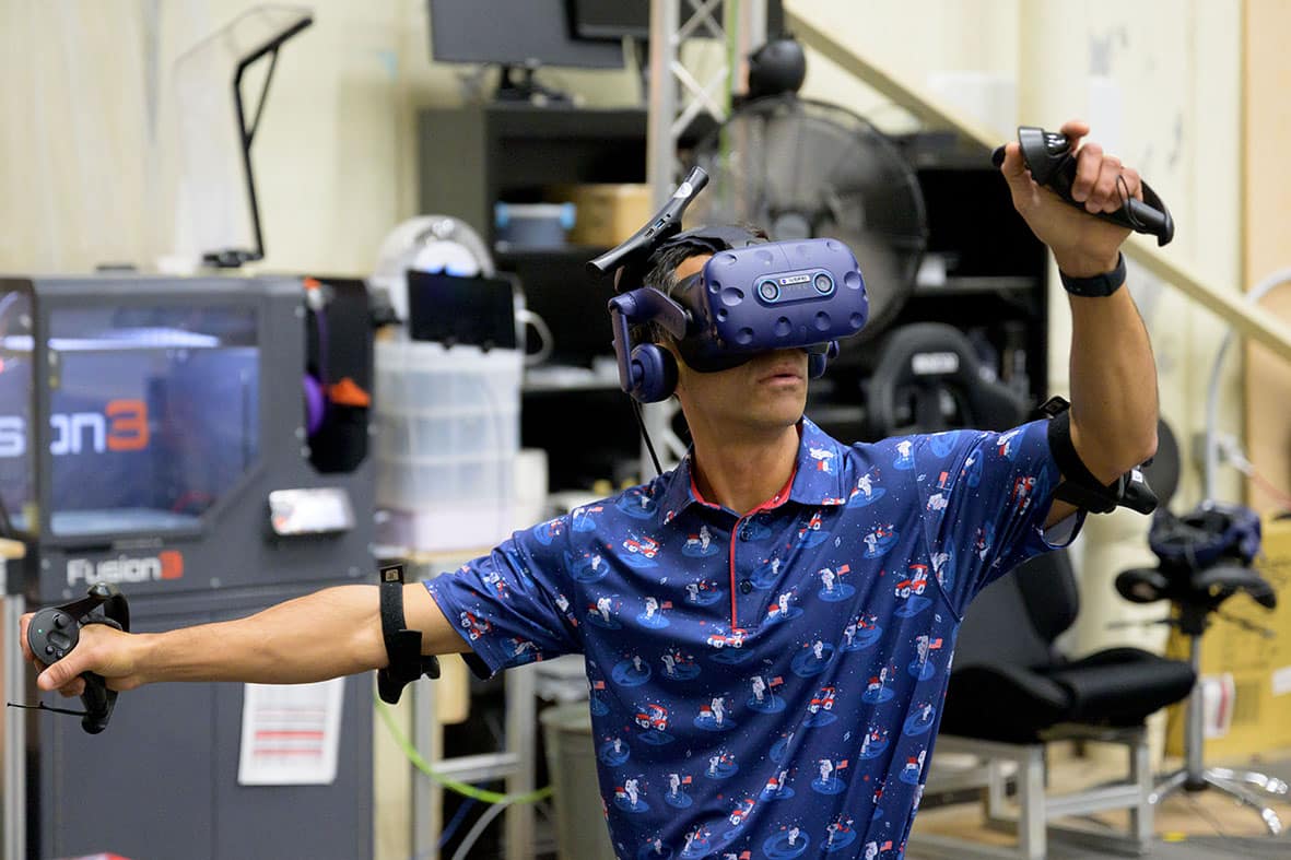 Person wearing a virtual reality headset and motion sensors using handheld controllers in a tech lab environment.