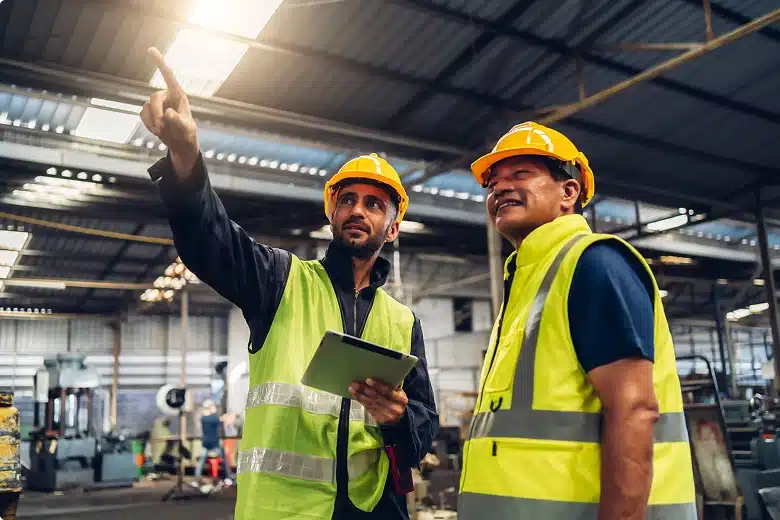 Two engineers in safety vests and helmets reviewing operations inside a large industrial facility with a tablet.