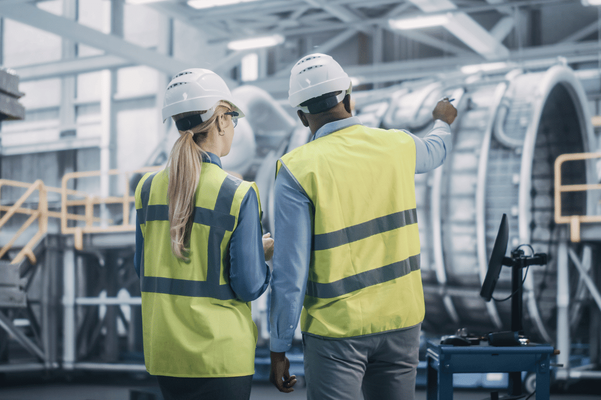 Two engineers in high-visibility vests and hard hats inspect large aerospace components inside a modern industrial facility, representing quality control and precision engineering.