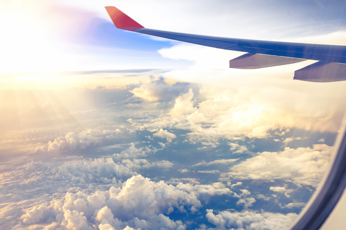 Scenic view of fluffy clouds and soft sunlight through an airplane window with a visible wing, capturing the beauty of air travel at high altitude.