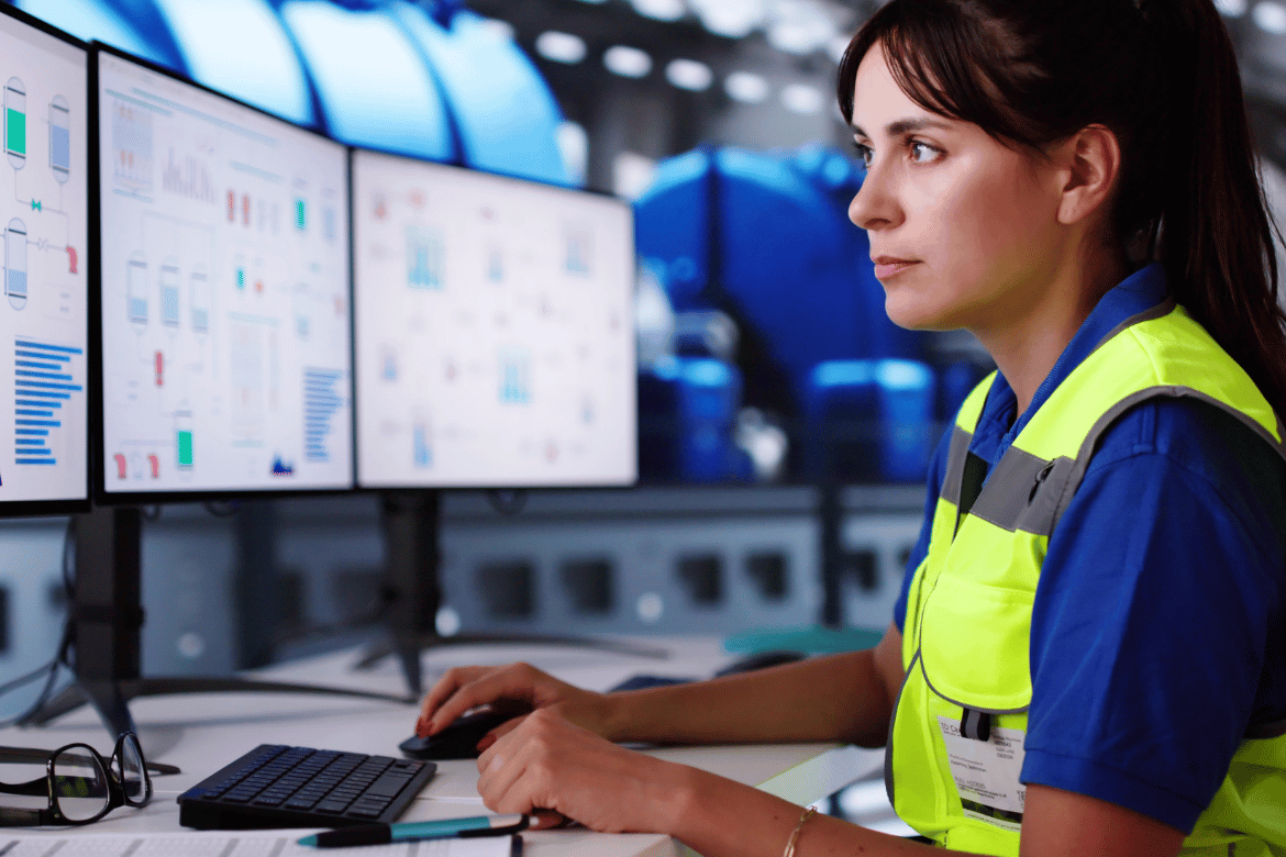 Female engineer in high-visibility vest working at a multi-monitor control station, analysing industrial system data and performance metrics in a high-tech environment.