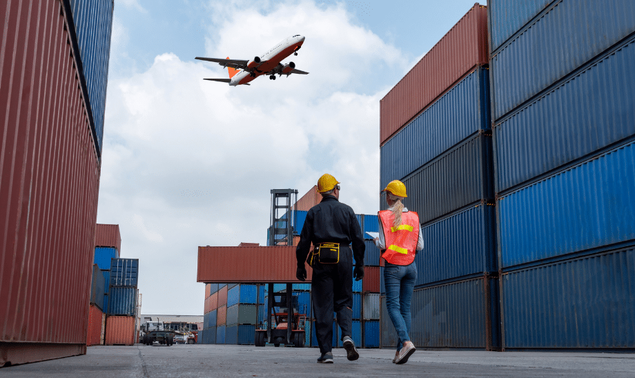 Two workers in safety gear walk between stacked shipping containers while a cargo plane flies above, representing global logistics and freight transport.