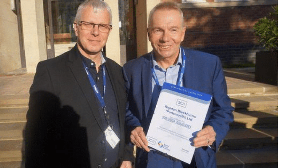 Two people smiling and holding an SC21 Silver Award certificate standing outside a building in sunlight