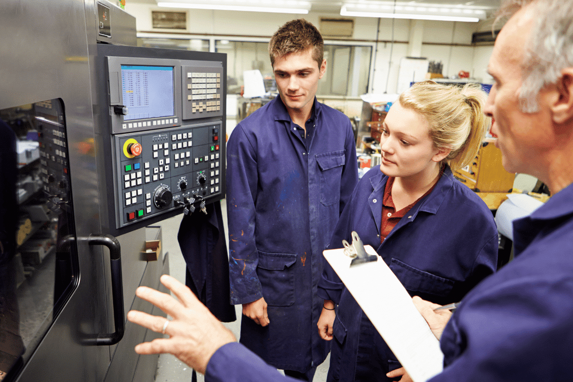 Young apprentices in blue overalls observe a supervisor explaining CNC machine controls in a workshop, representing hands-on engineering training and skills development.